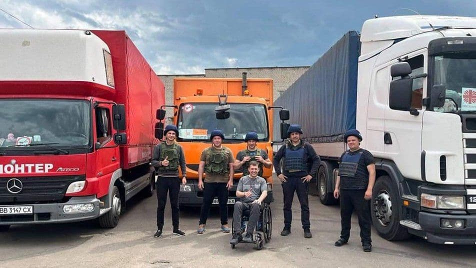 A group of men in military vests and helmets posing in front of three large trucks—red, orange, and white. One man is sitting in a wheelchair, and everyone appears smiling.