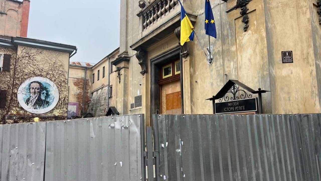 Entrance to a historic building—the Museum of the History of Religion in Lviv. Above the doorway is a sign with the museum’s name, flanked by the Ukrainian and European Union flags. A metal fence partially blocks the entrance, indicating renovation work. On the left is a mural of Nicolaus Copernicus on a vine-covered wall. Nearby are plaques and signs, including a street sign reading “1 Muzeina Street.” The scene reflects cultural heritage and public accessibility.
