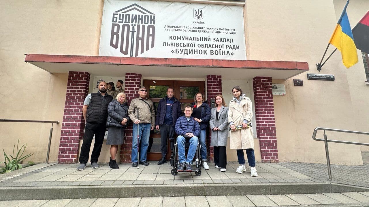 A group of people standing in front of a building with a sign above the entrance that reads: "House of Warriors, Ukraine, Department of Social Protection of the Population of Lviv Region State Administration, Communal Institution of Lviv Regional Council 'House of Warriors'." There are two flags on the right side—one of Ukraine and another with red and black colors. The building’s address is marked as "1a." The group includes a person in a wheelchair and others standing around, indicating a communal establishment related to social protection services in Lviv, Ukraine, specifically for veterans.