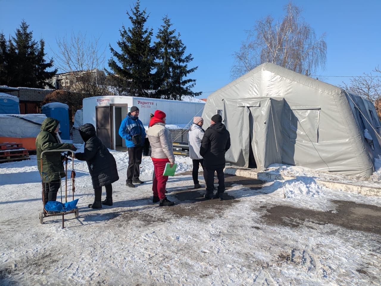 People gathered near a large humanitarian aid tent located in a snowy area. In the background, a mobile shelter labeled “Shelter” and portable toilets. Two elderly women in winter clothing are talking, one leaning on a walker.