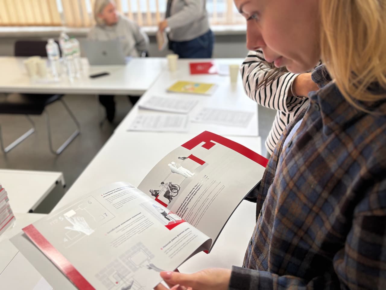 Учасниця читає розділ “Альбому безбар’єрних рішень”, розглядаючи ілюстрації та технічні схеми доступності.»