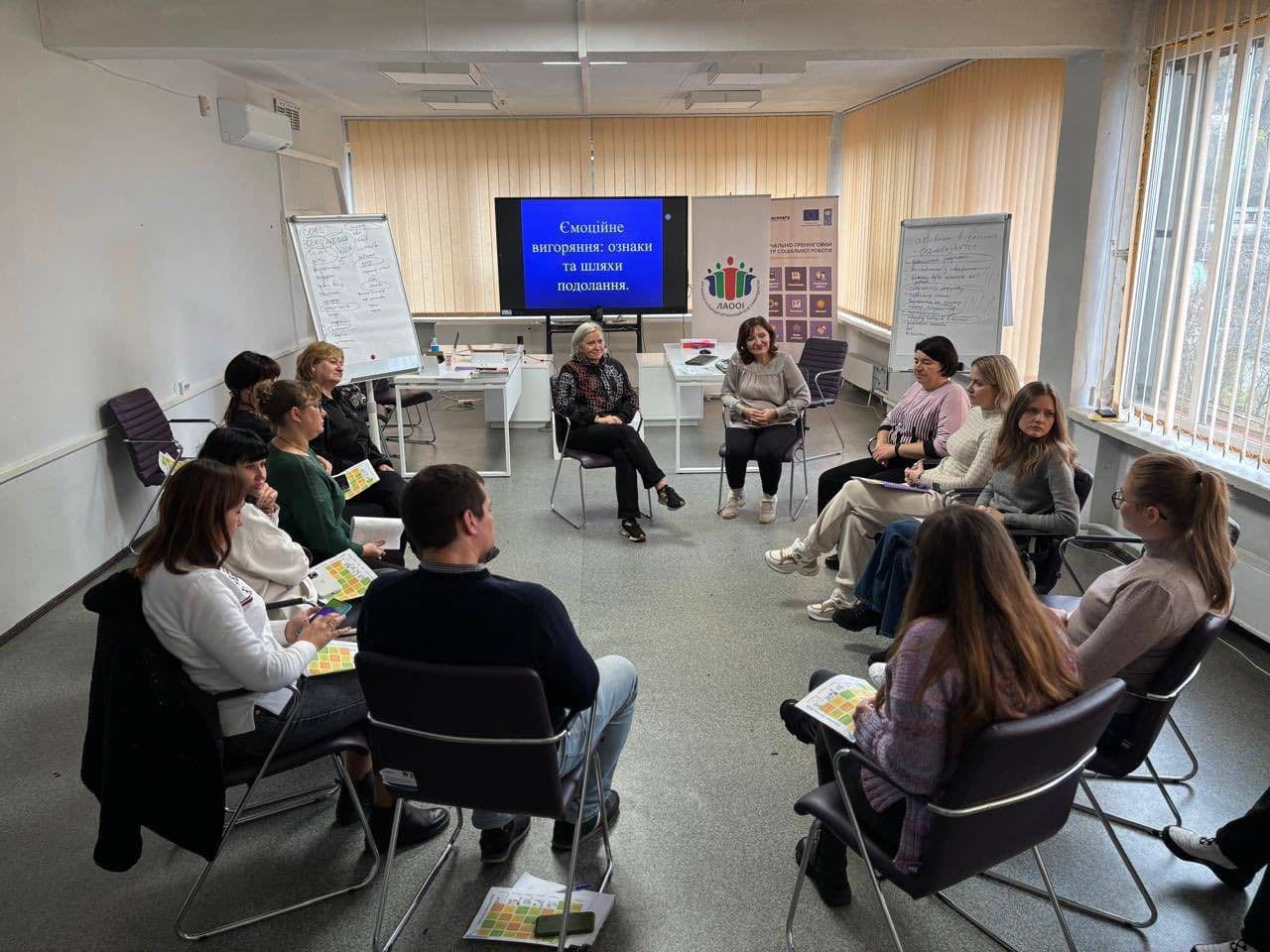 A group of 14 people sits in a circle in a brightly lit room, participating in a training session. Some hold colorful handouts. A presentation slide on the screen reads “Emotional burnout: signs and ways to overcome it.” Behind them are two flip charts with notes on social support, rest, physical activity, hobbies, and psychological help. The setting is professional and educational.