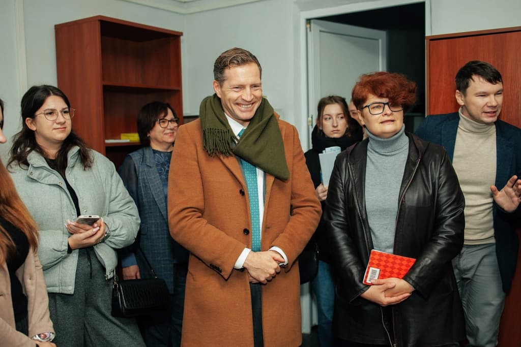A group of eight people stands in an indoor space resembling an office or meeting room. At the center, a smiling man wears a brown coat, green scarf, and blue tie. Others hold notebooks, folders, or a smartphone. In the background are wooden cabinets and an open door. The scene captures a moment of professional interaction or inspection.