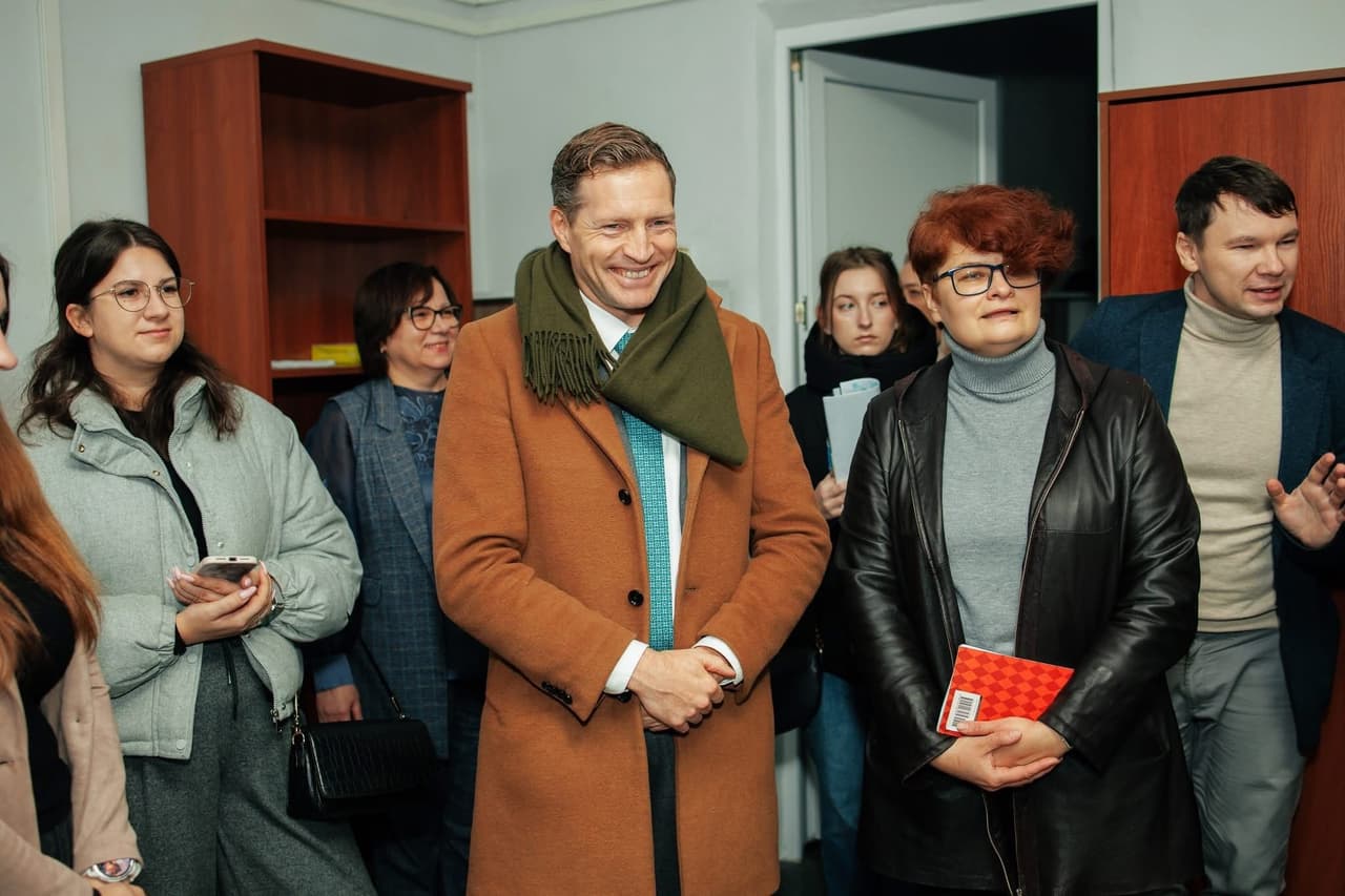 A group of eight people stands in an indoor space resembling an office or meeting room. At the center, a smiling man wears a brown coat, green scarf, and blue tie. Others hold notebooks, folders, or a smartphone. In the background are wooden cabinets and an open door. The scene captures a moment of professional interaction or inspection.