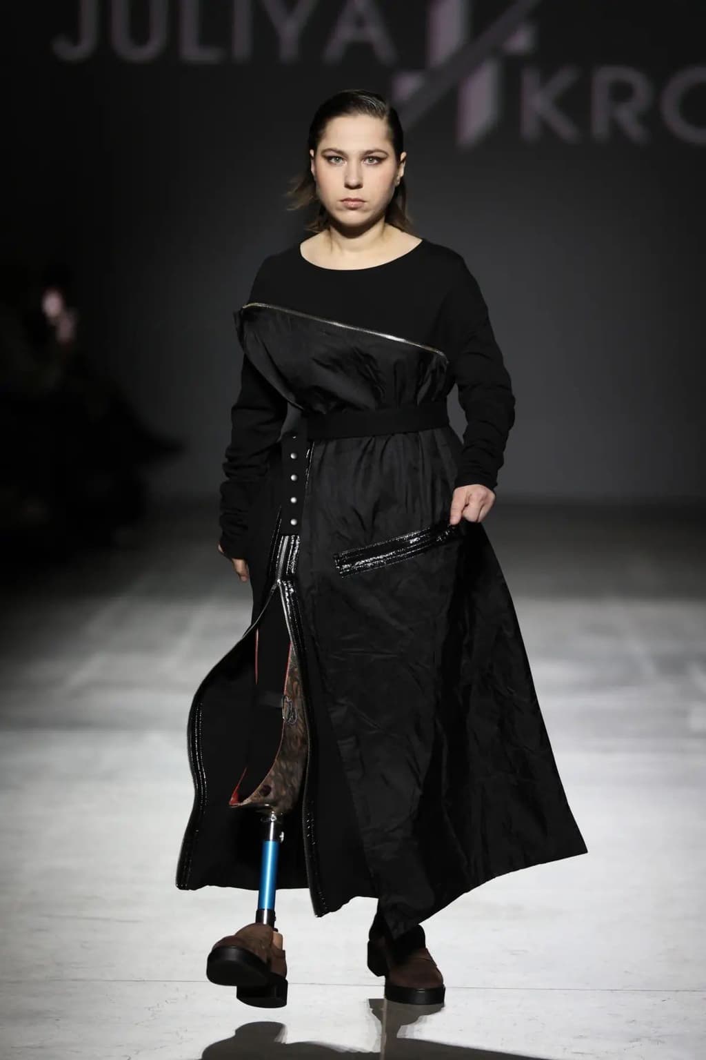 A fashion model walks the runway wearing a black asymmetrical dress with a diagonal zipper and shiny accents. The slit reveals a prosthetic leg with a blue metal rod and patterned socket. The model wears brown platform shoes. Behind her, the brand name “JULIYA KROS” is projected on the wall. The scene highlights inclusivity and diversity in fashion, showcasing adaptive design and representation of individuals with disabilities.
