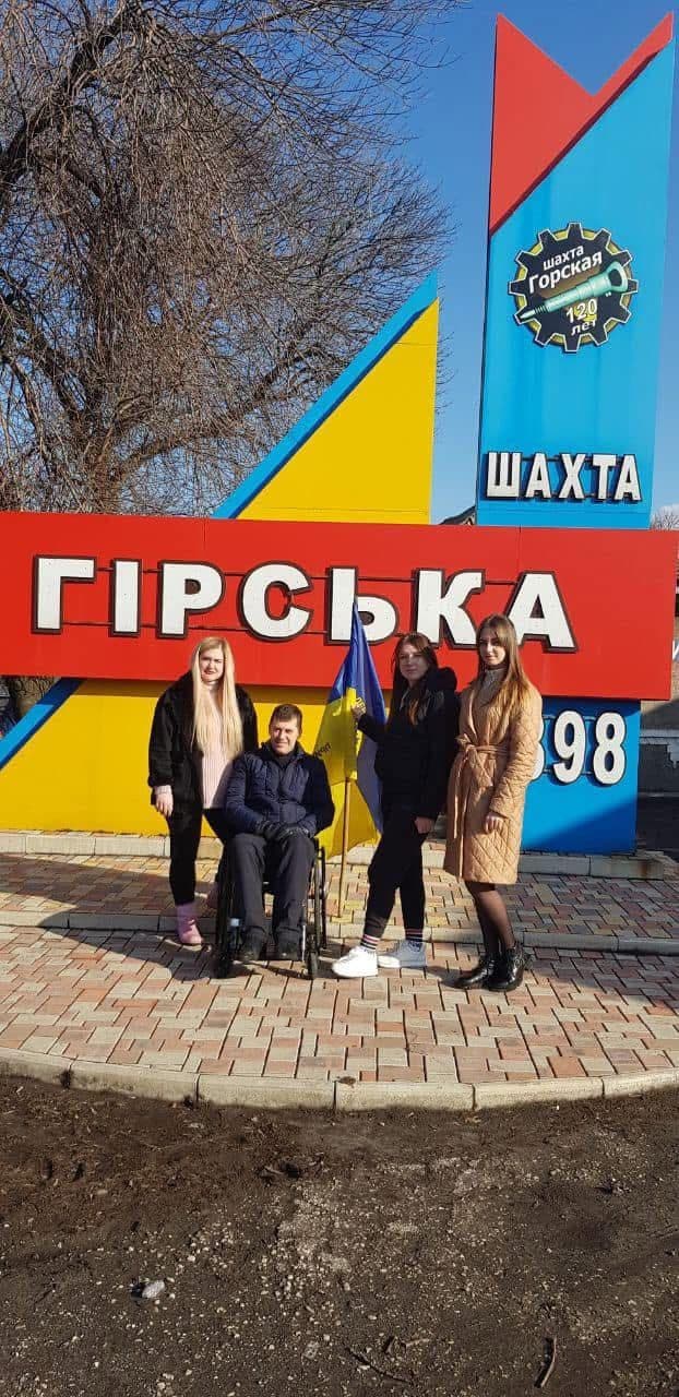 Чотири людини позують перед яскравим пам’ятником із кольоровими панелями — червоною, жовтою та синьою. Один із учасників сидить в інвалідному візку, інший тримає український прапор. На червоній панелі великими білими літерами написано «ГІРСЬКА». На синій панелі — емблема у формі шестерні з написом «шахта горская» та «120 лет», що позначає ювілей шахти. Нижче великими літерами — «ШАХТА», а також рік «1898». Сцена передає вшанування історії та спадщини Гірської шахти.