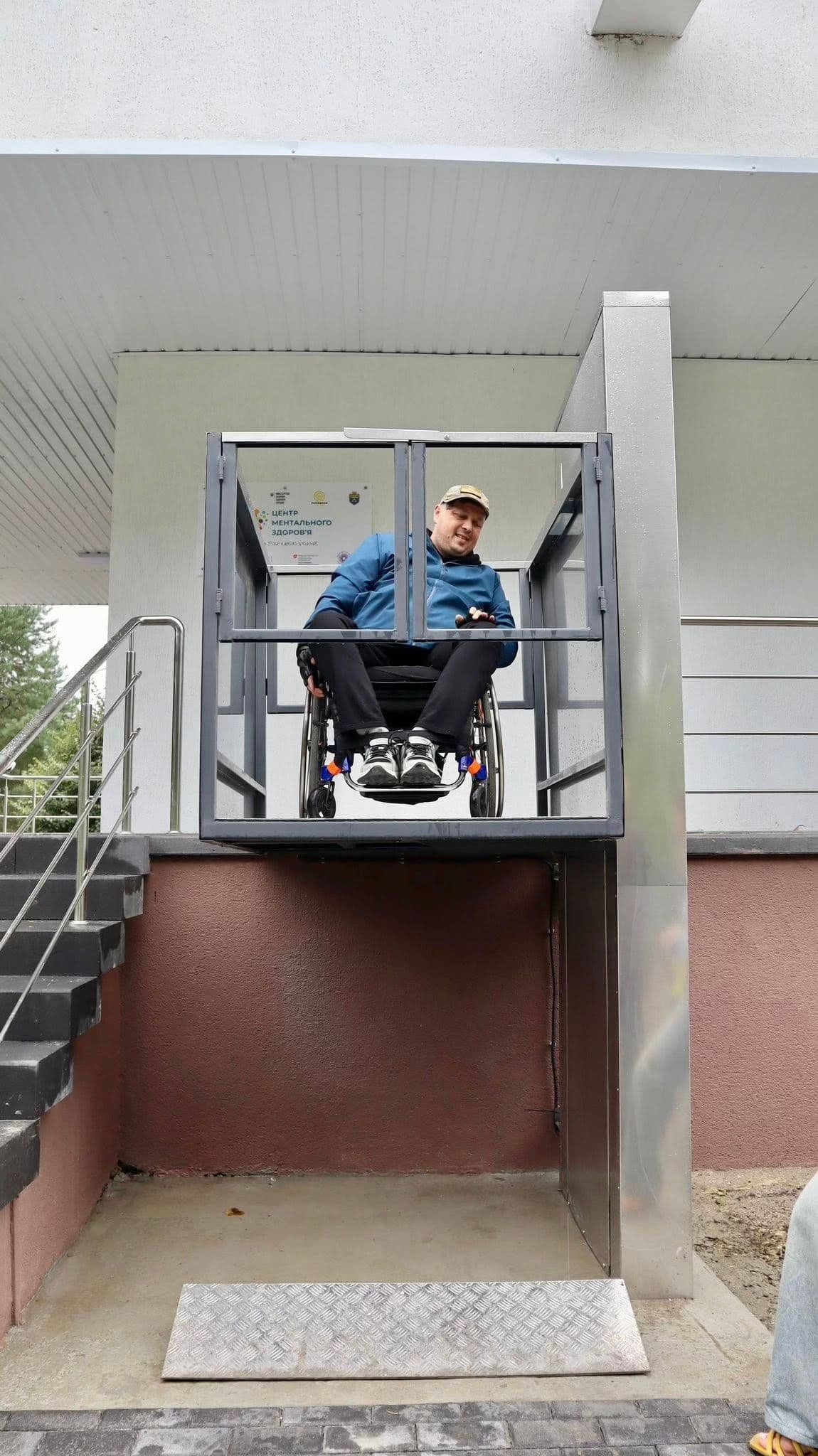 Людина у візку користується вертикальним платформним підйомником, щоб піднятися на вищий рівень, ймовірно, до входу в будівлю. Підйомник обладнаний захисними бар’єрами та розташований поруч зі сходами, забезпечуючи доступну альтернативу для осіб з порушеннями мобільності. Людина на платформі розміщена безпечно. Сцена демонструє інклюзивну інфраструктуру, що сприяє самостійності та рівному доступу.