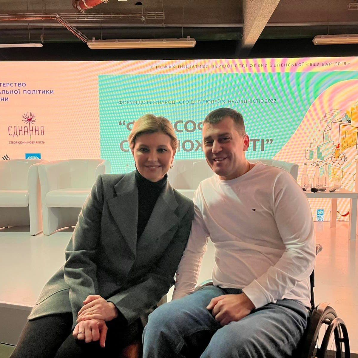 A woman sits next to a man in a wheelchair in a hall, with an image displaying the conference topic on a turquoise background projected on the wall.