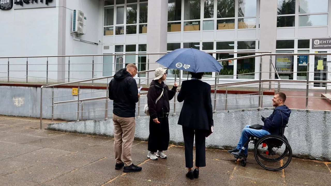 Четверо людей перебувають біля будівлі з великими вікнами та пандусом, що веде до входу. Одна особа у візку знаходиться біля основи пандуса, троє інших стоять поруч і спілкуються. Один із них тримає парасолю. На фасаді — вивіска кирилицею, що вказує на російськомовний регіон. Сцена акцентує увагу на інклюзивній інфраструктурі та доступності громадських просторів.