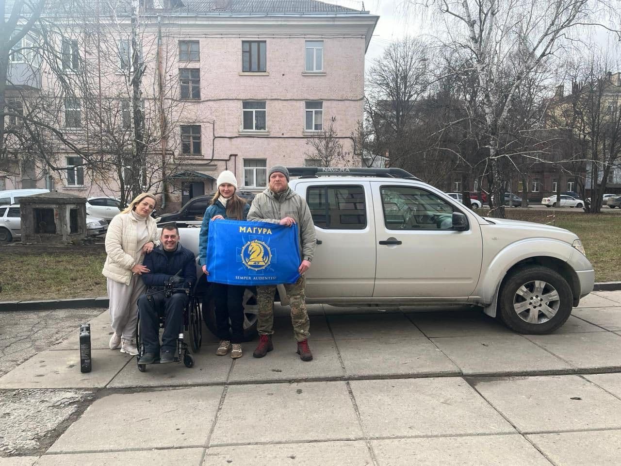 Four individuals stand beside a silver Nissan Navara pickup truck parked on a paved area near a residential building. One person is seated in a wheelchair, while the other three stand, two of them holding a blue flag with a yellow emblem and the words “МАГУРА” and “SEMPER AUDENTES.” The emblem features a stylized horse head and shield. In the background are a pink building, leafless trees, and parked vehicles. The scene captures a moment of solidarity or support, possibly linked to a community or veteran organization.