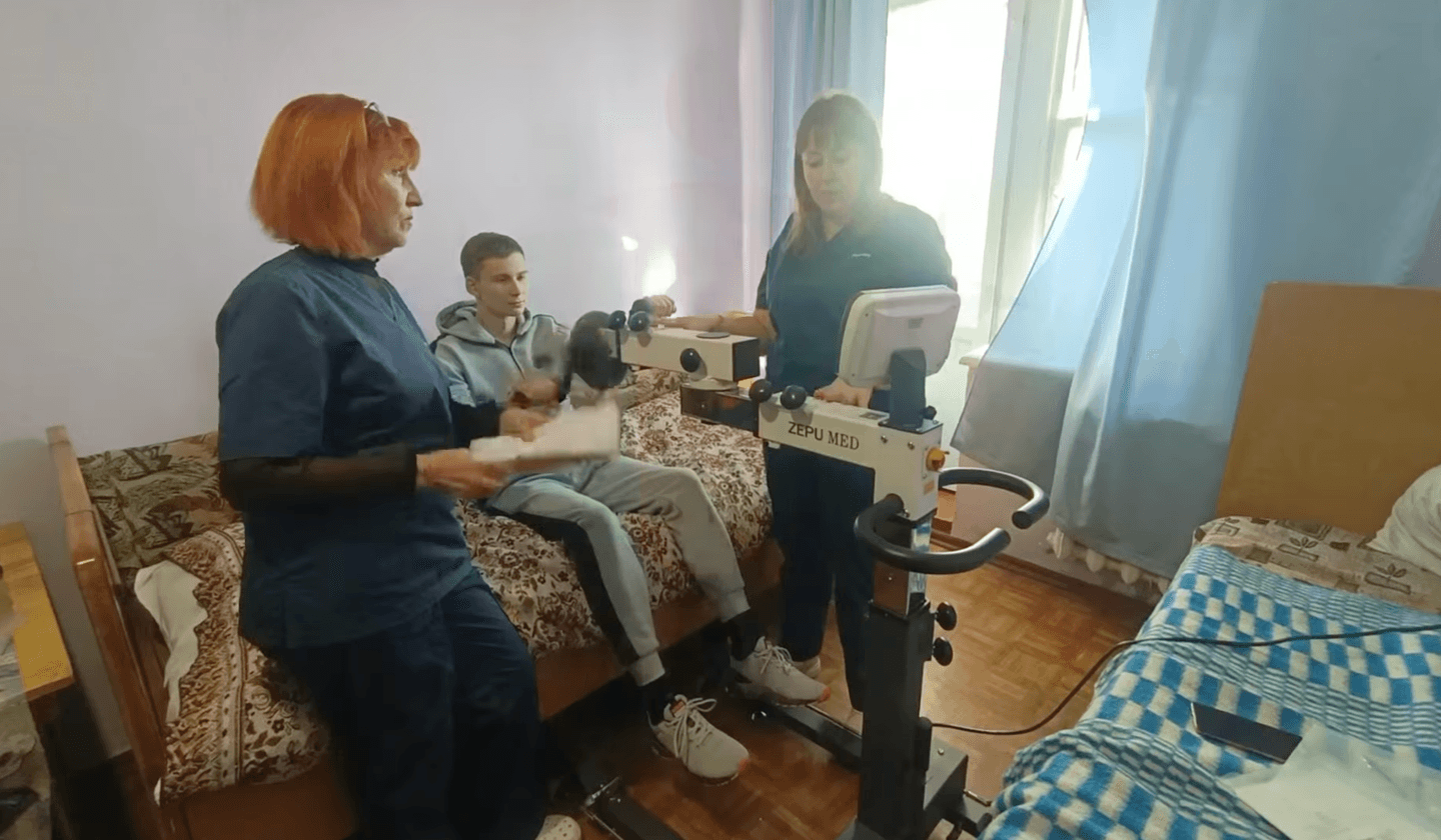 A patient in a gray tracksuit sits on a bed holding the handles of a ZEPUMED rehabilitation machine. Two nurses in blue uniforms assist in setting up the equipment. The room has wooden flooring, and blue curtains are near the window.