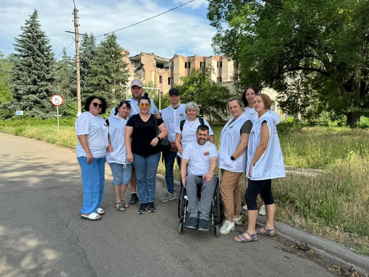 A group of volunteers in white uniforms standing on a street with trees and a ruined building in the background. A man in a wheelchair is in the center.