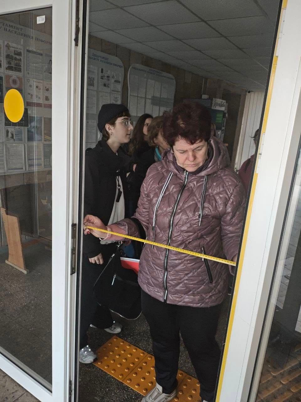 A group of people stands at a building entrance. One person in a purple jacket holds a yellow measuring tape across the doorway. The glass doors feature a yellow sticker, and tactile paving is visible on the floor. Informational boards are mounted on the wall. The scene suggests an accessibility inspection or compliance check.