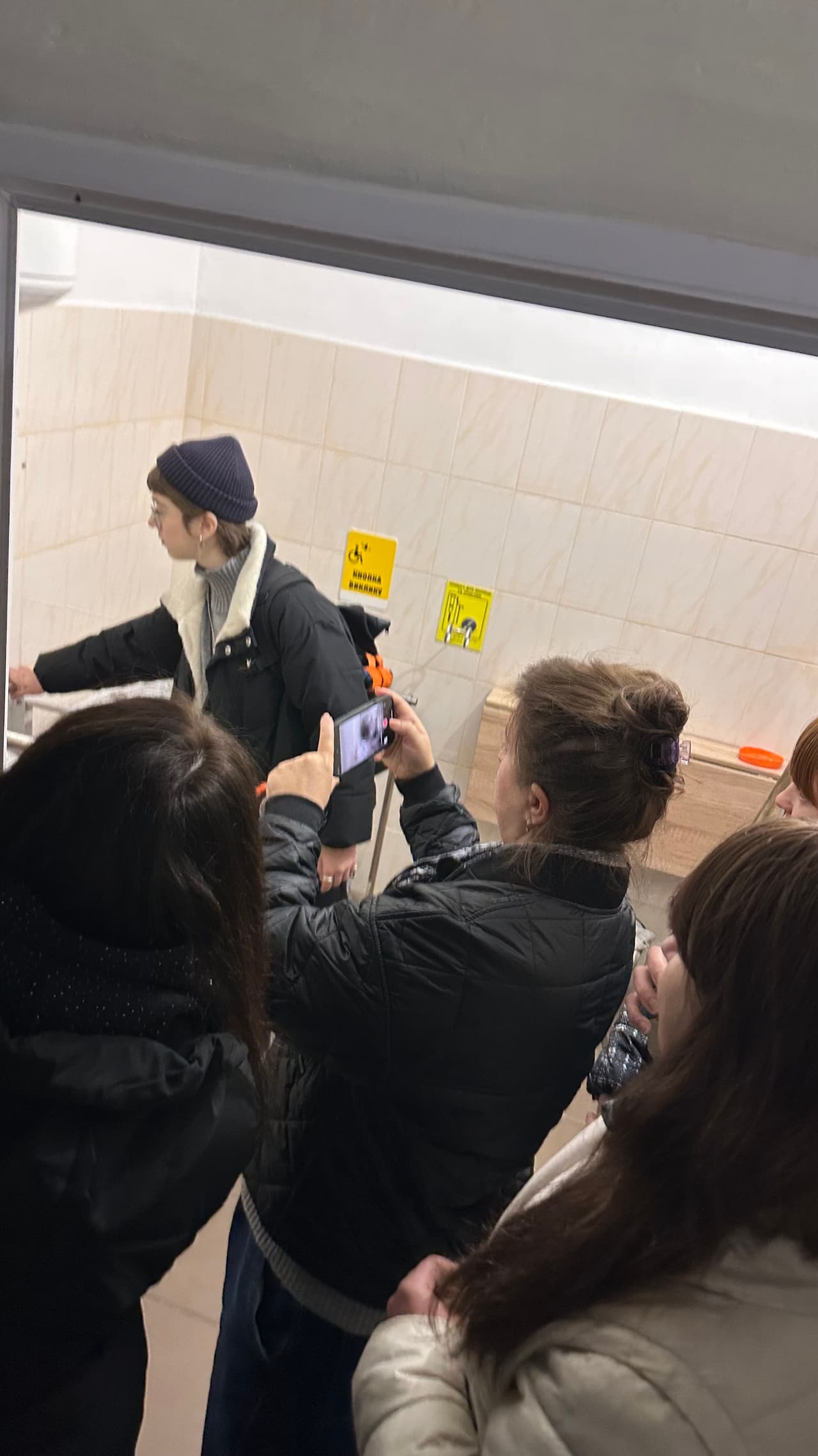 Група учасниць проводить огляд санітарно-гігієнічного приміщення під час практичного заняття з оцінки доступності. На стіні видно інклюзивні піктограми, поручні та сантехнічне обладнання. Одна з учасниць фіксує елементи приміщення на телефон.»