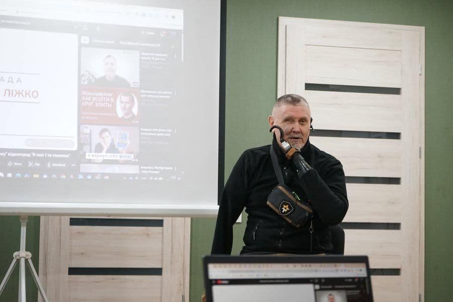 На зображенні чоловік із протезом руки виступає перед аудиторією. Він одягнений у чорний одяг і має невелику сумку через плече з емблемою у вигляді зірки. Позаду нього на екрані проєктується інтерфейс YouTube з кількома відео та написами кирилицею, серед яких видно заголовки «РАДА ЛІЖКО», «Малкович: КАК ВОЙТИ В КРУГ ЭЛИТЫ» та «вирусный блогер». На передньому плані видно ноутбук, екран якого також відображає YouTube. Атмосфера нагадує лекцію або презентацію, пов’язану з медіа чи технологіями.