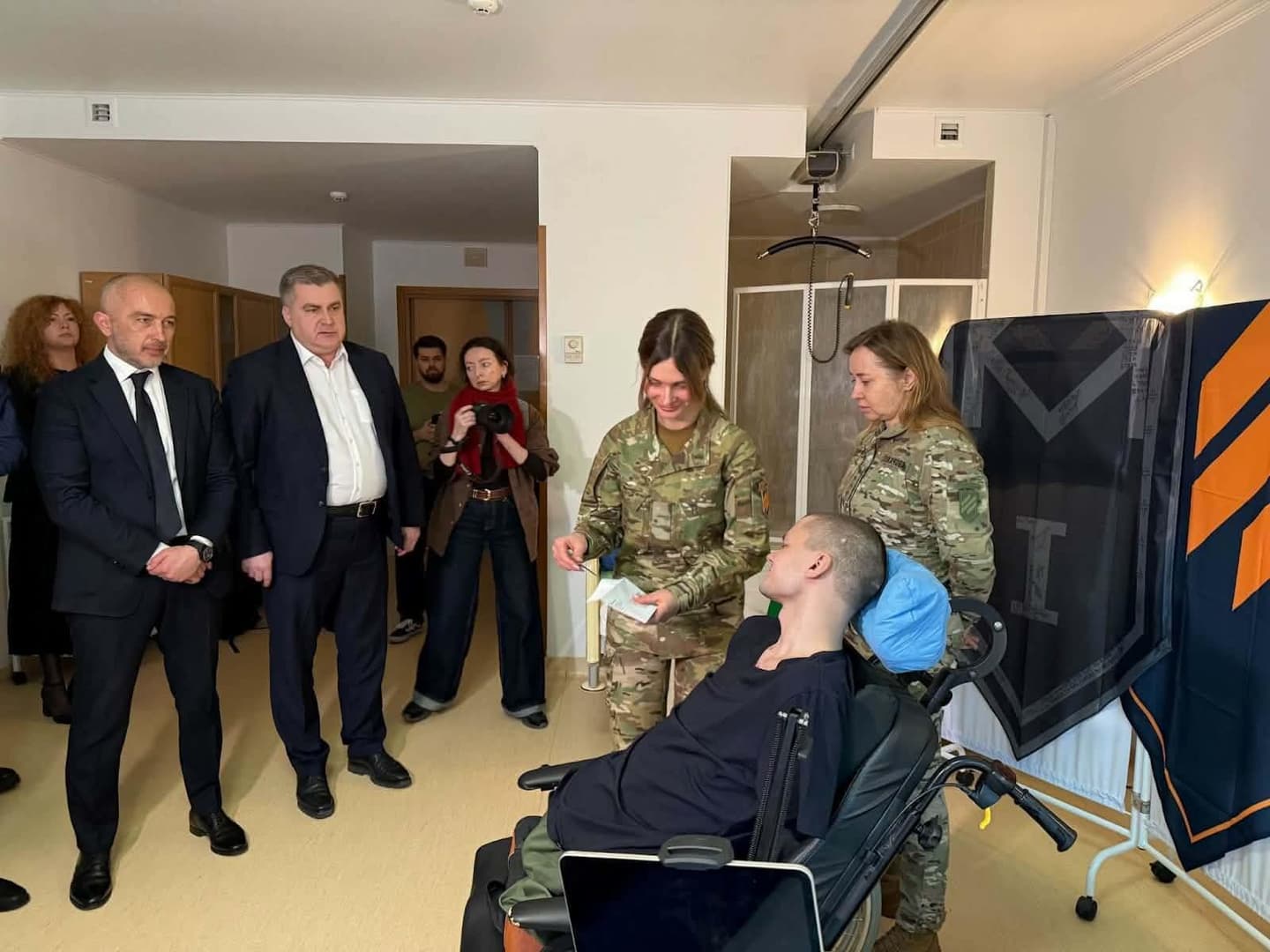 A group of people in a room, including military personnel in camouflage and individuals in formal attire. At the center, a man in a wheelchair interacts with two soldiers, one smiling and handing him an item. In the background, observers include a man in a suit and a woman with a camera. The room resembles a medical or rehabilitation facility, with a ceiling lift, wheelchair, and colorful geometric banners. The scene reflects a moment of recognition, support, or an official visit.