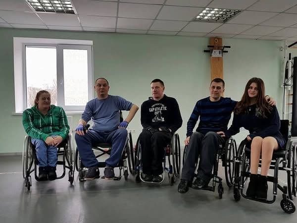 Five people are sitting in wheelchairs in a room that looks like a rehabilitation center. They appear friendly and relaxed, with some touching each other or smiling. The room contains exercise equipment, and a large window behind them lets in natural light. They are all dressed in comfortable casual clothing, and their facial expressions convey positive emotions.