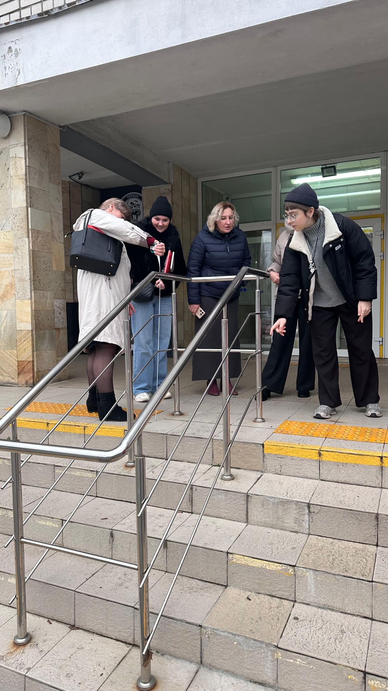 Група учасниць тренінгу оглядає вхідні сходи будівлі під час практичного заняття з оцінки доступності. На фото видно тактильну плитку, поручні та вхід до установи.» EN — Alt-text