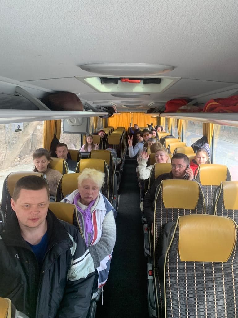 На зображенні видно інтер'єр автобуса з кількома пасажирами, які сидять на своїх місцях. Сидіння обтягнуті матеріалом з жовто-чорною кліткою, а на вікнах автобуса висять жовті штори. Деякі пасажири дивляться вперед, інші зайняті розмовою або жестами. У верхніх відсіках розміщені деякі багажі. Автобус, здається, рухається, оскільки вид за вікнами показує розмитий пейзаж. Це зображення цікаве тим, що показує момент подорожі, підкреслюючи розташування і взаємодію пасажирів в автобусі.