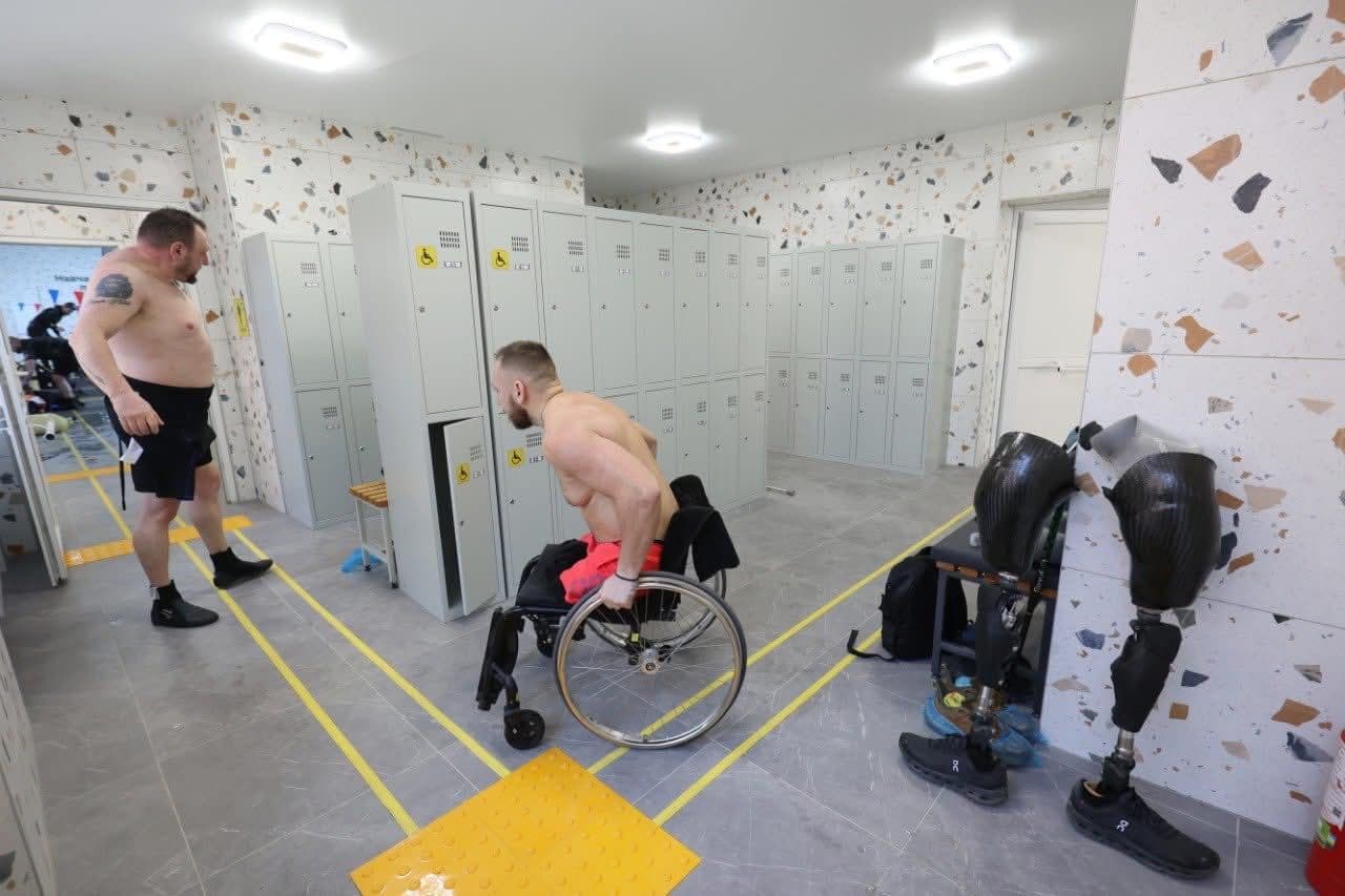 Роздягальня з кількома людьми, один із яких користується інвалідним візком. Він без сорочки та відкриває шафку, позначену жовтим символом доступності. Поруч стоїть інший чоловік без сорочки. Справа на підлозі біля лавки лежать дві протези ніг із чорним взуттям. У приміщенні є кілька шафок, лавка, плиткова підлога з жовтою тактильною плиткою та напрямними лініями. Стіни мають оздоблення у стилі терраццо, освітлення забезпечують стельові світильники. Сцена демонструє приклад доступності та адаптивного обладнання у громадських просторах.