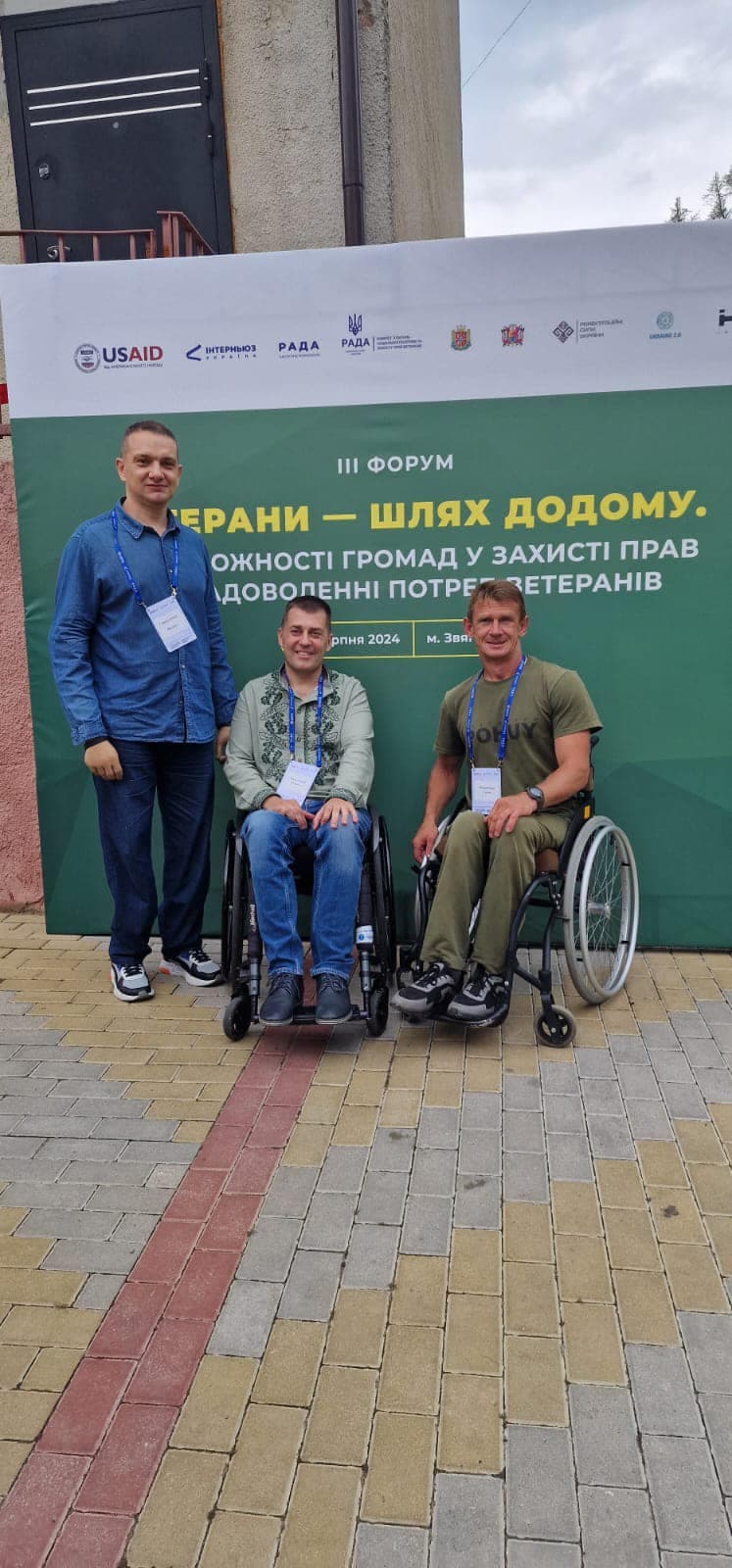 Чоловік стоїть поруч із двома чоловіками, які сидять в інвалідних візках, на фоні зеленого банера.
