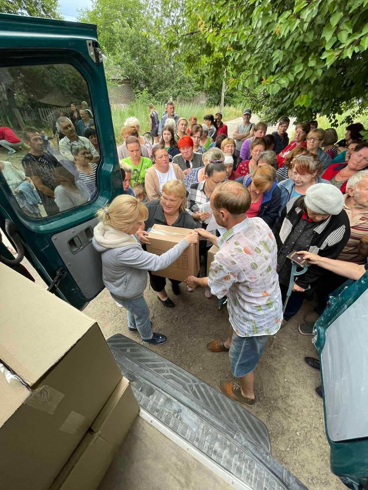 People distributing humanitarian aid hand out boxes of food and essential items to a large group of local residents standing in line near a green van. Some individuals are holding documents while waiting for their turn.