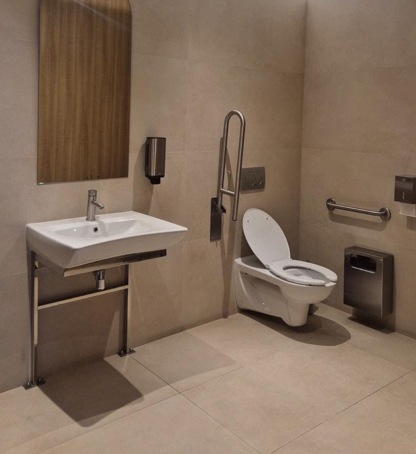 A modern public restroom designed for accessibility. It features a wall-mounted toilet with a raised seat, a foldable support rail beside it, and a horizontal grab bar on the adjacent wall. A wall-mounted sink with wheelchair clearance is equipped with a soap dispenser and a large vertical mirror above. A toilet paper holder and waste bin are also present. The interior has beige tiles and stainless steel fixtures, emphasizing functionality and inclusive design.