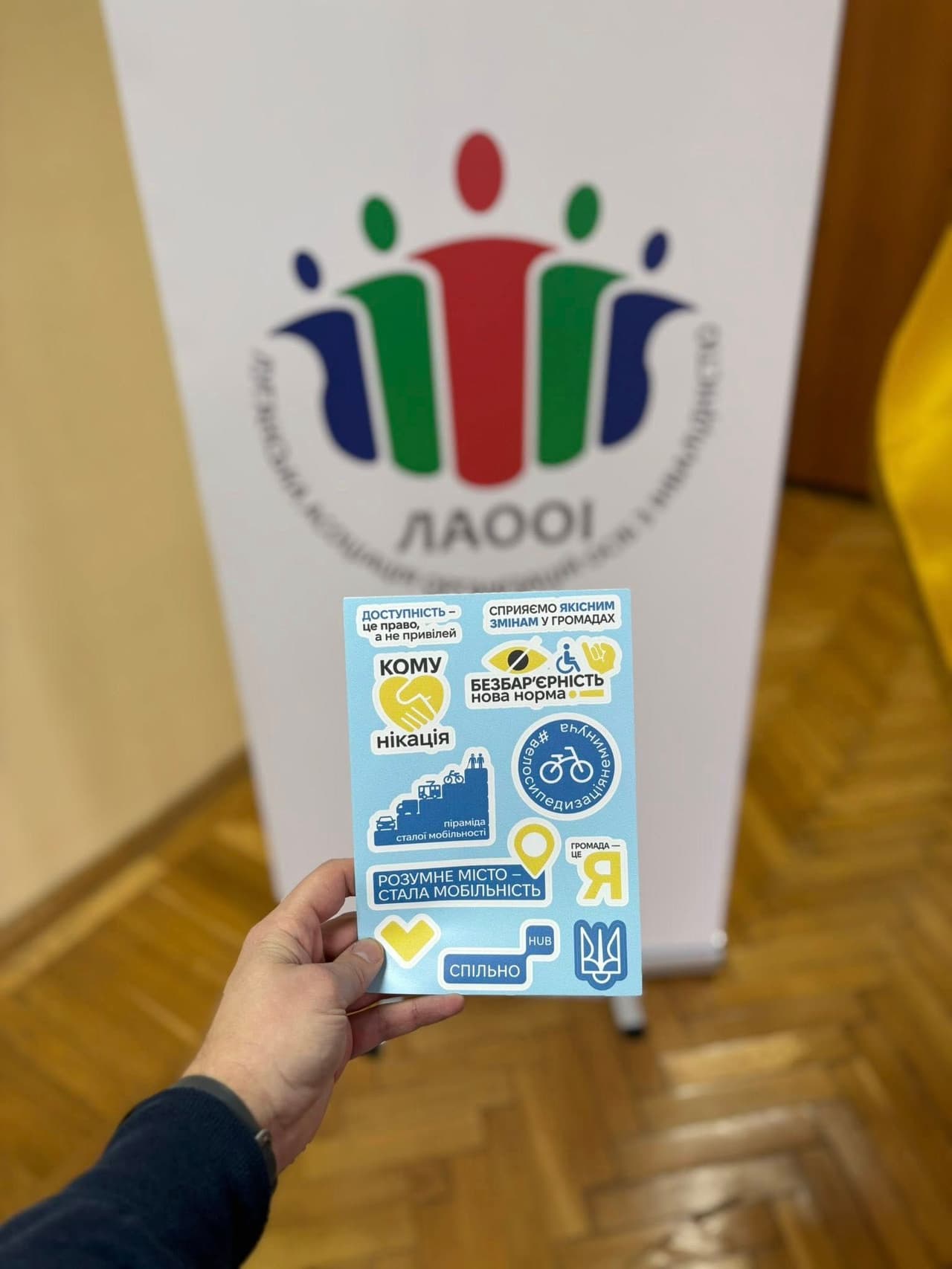 A person holds a sheet of stickers in front of a banner featuring the logo of the Lviv Association of Persons with Disabilities (LAOOI): stylized human figures in red, green, and blue. The stickers display Ukrainian phrases promoting accessibility, mobility, barrier-free environments, and civic inclusion, such as “Accessibility is a right, not a privilege” and “Community is me.” The scene conveys messages of inclusion and advocacy.