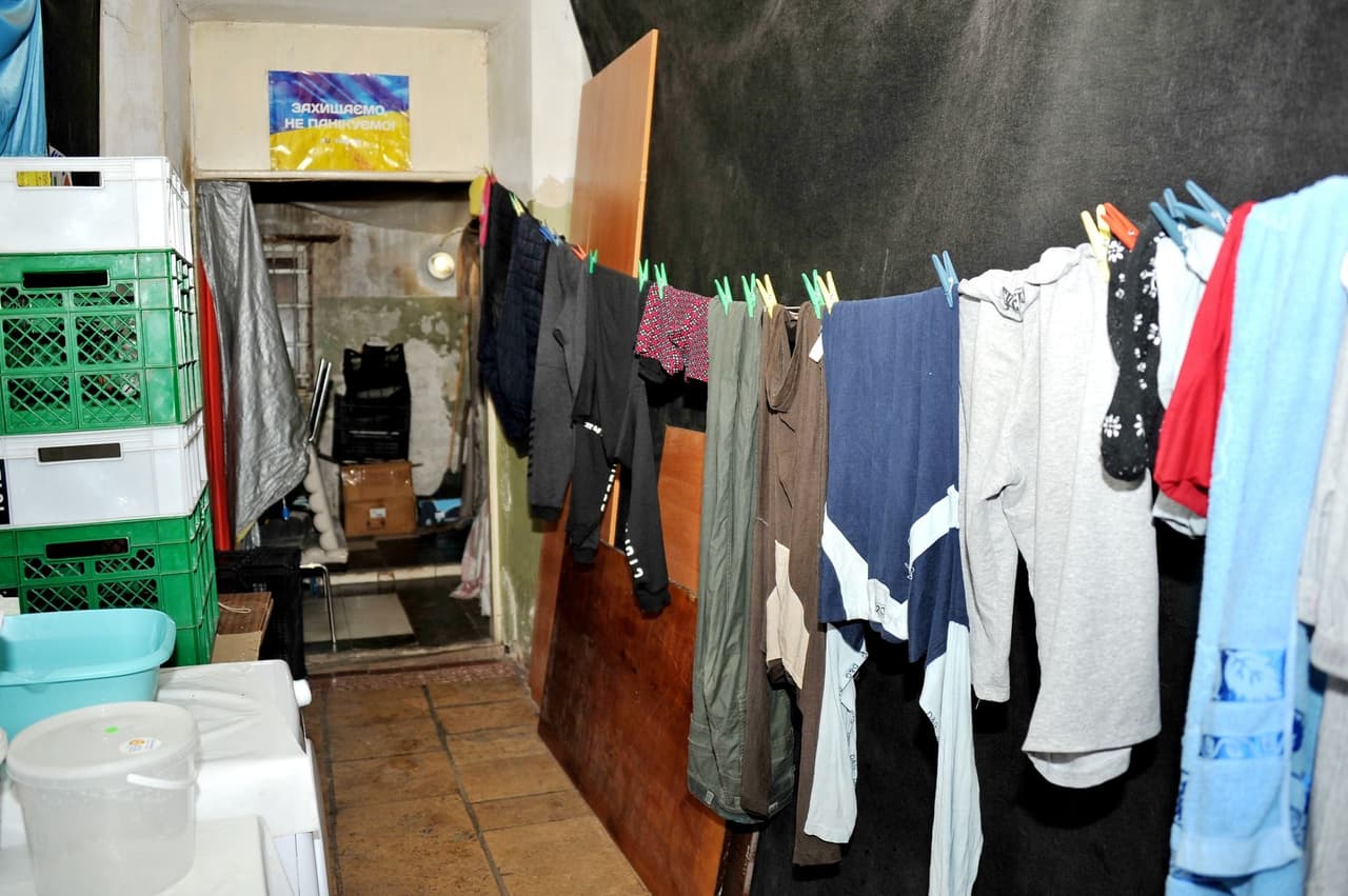 A clothesline with hanging laundry stretches across the room. On the left, plastic crates and kitchenware are visible, while boxes and belongings are stored in the background. A sign on the wall reads “Protect, don’t panic!”.