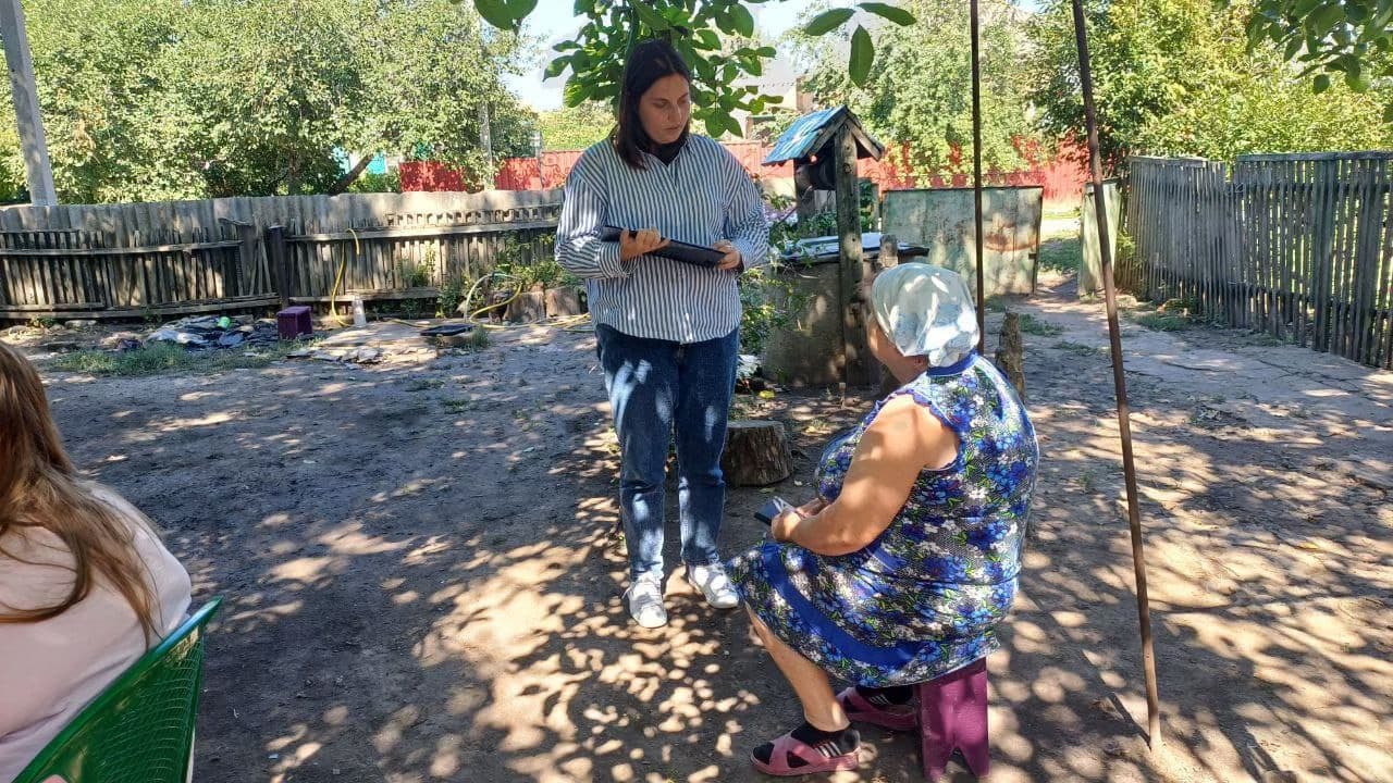 Three individuals are gathered in a backyard or garden area. One person in a striped shirt and jeans stands and appears to be speaking to a seated woman in a blue floral dress and white headscarf, who sits on a small stool. A third person, partially visible on the left, has long hair and sits on a green chair. The background includes a wooden fence, a covered well, and scattered household items such as buckets and containers. Trees and greenery cast shadows across the ground, suggesting a warm, informal outdoor conversation in a rural setting.