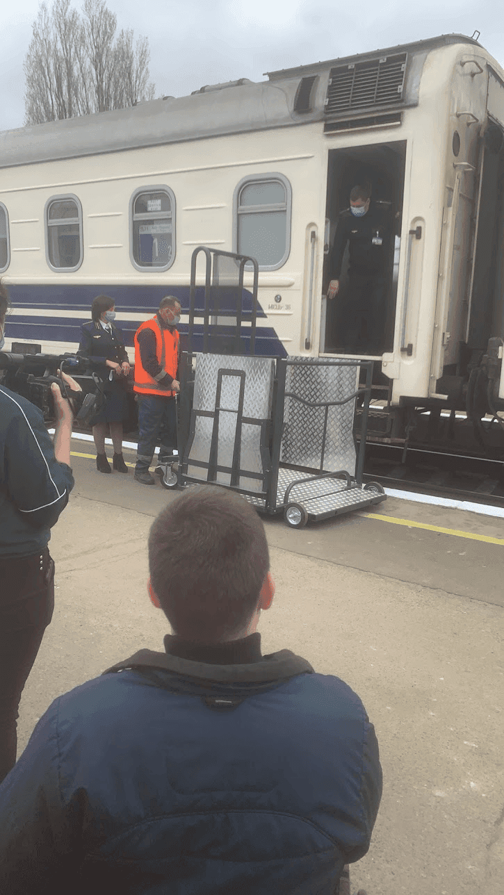 Потяг із біло-блакитним екстер'єром стоїть на платформі. Декілька людей знаходяться поруч, серед них працівник у помаранчевому жилеті безпеки, який керує металевим візком. Інша особа в уніформі, можливо, провідник або працівник станції, стоїть неподалік. Третя людина тримає камеру і знімає подію. Всередині потяга видно особу в масці та уніформі. Металевий візок, схоже, використовується для завантаження або розвантаження предметів, підкреслюючи активність на залізничній станції.