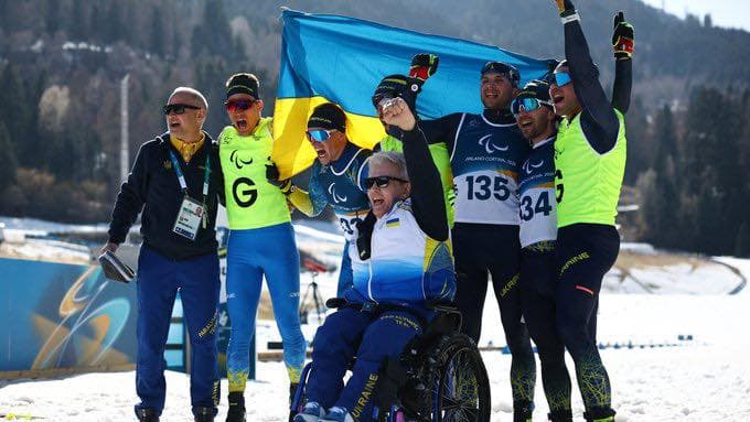 A group of Ukrainian para-athletes celebrate victory at a winter sports event. One athlete in a wheelchair holds the Ukrainian flag, while others cheer and raise their arms. Their uniforms display the word “Ukraine” and the Paralympic symbol, with bib numbers “135” and “34” visible. The snowy mountain landscape forms the backdrop. The scene captures triumph, unity, and resilience of Ukrainian Paralympians.