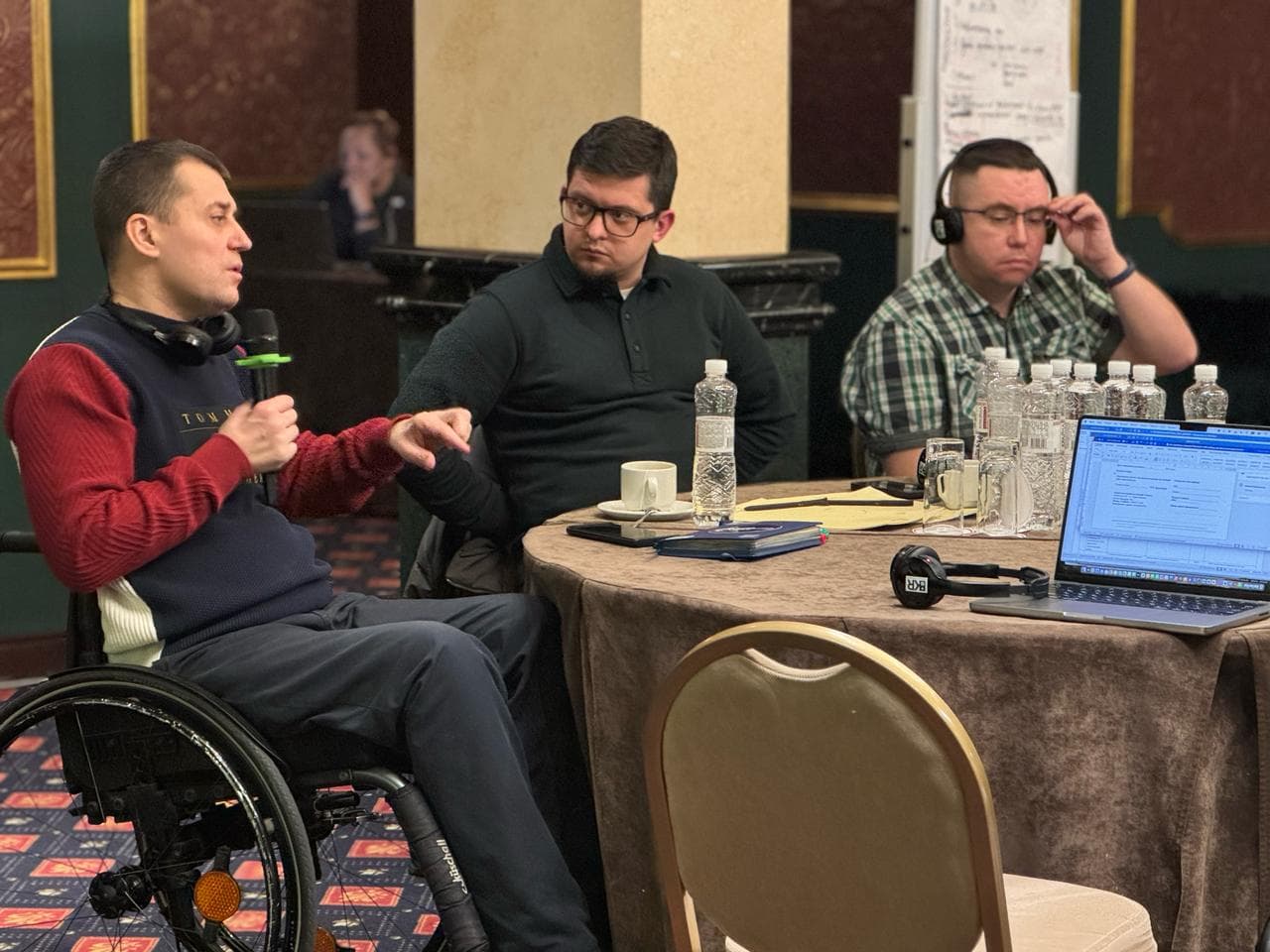 Conference participants sit around a round table, attentively listening to the speaker. A laptop and projector are in the foreground.