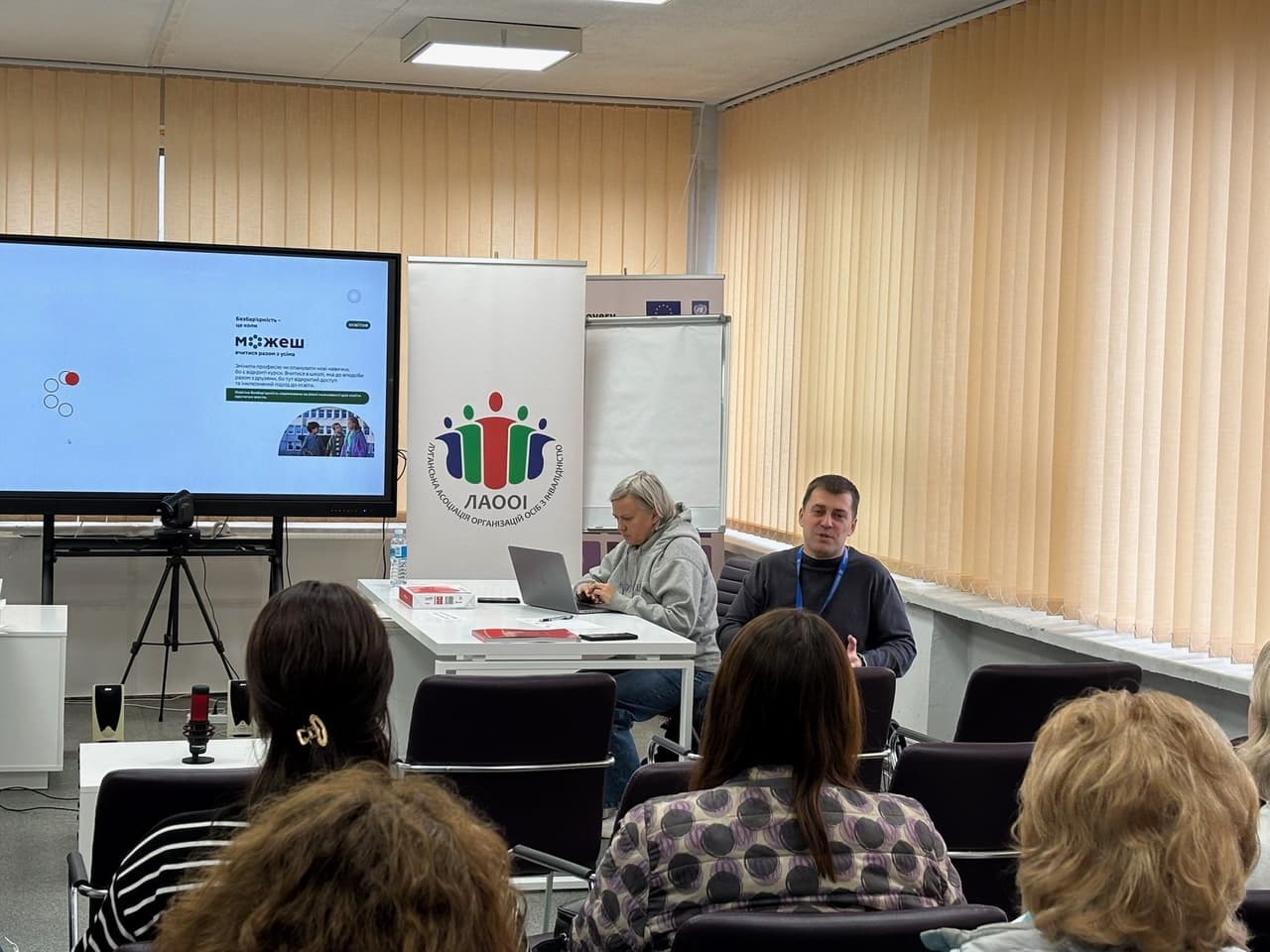 Семінар у конференц-залі: учасники сидять перед доповідачем, який виступає поруч із ноутбуком. На екрані — сайт «Можеш» із текстом про благодійність і підтримку постраждалих від війни. Попереду встановлена камера на тринозі. Позаду — банер Луганської асоціації організацій осіб з інвалідністю.