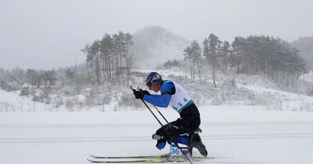 A para-athlete competes in a cross-country skiing event on a snowy mountain course. The athlete uses a sit-ski, a specialized sled for individuals with lower limb impairments. Dressed in a blue and white racing suit, the athlete’s bib and equipment display the number “36.” Snow-covered trees and hills form the serene winter background. The scene highlights strength, resilience, and inclusivity in sports.