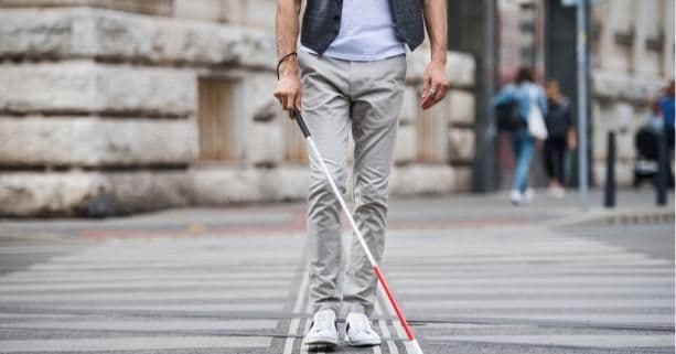 A person walks across a pedestrian crosswalk using a white cane, indicating visual impairment. They wear light-colored pants, a white shirt, and a dark vest. In the background, a stone building and several pedestrians on the sidewalk suggest an urban environment. The scene highlights independent mobility and accessibility for blind or visually impaired individuals in public spaces.