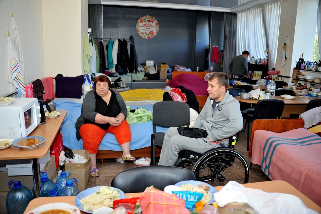 A man in a wheelchair and a woman are sitting in a room arranged as a temporary shelter. In the background, there are beds, personal belongings, a table with food, and other shelter residents.