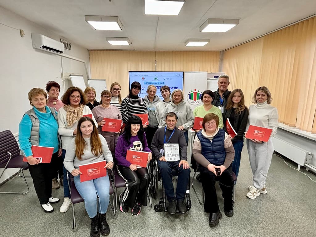 Group photo of participants in a brightly lit room, holding red folders labeled “Guide Without Barriers.” One person in the center holds a sign with the same title. A screen in the background displays Ukrainian logos and text, indicating an educational or advocacy event focused on accessibility and inclusion.