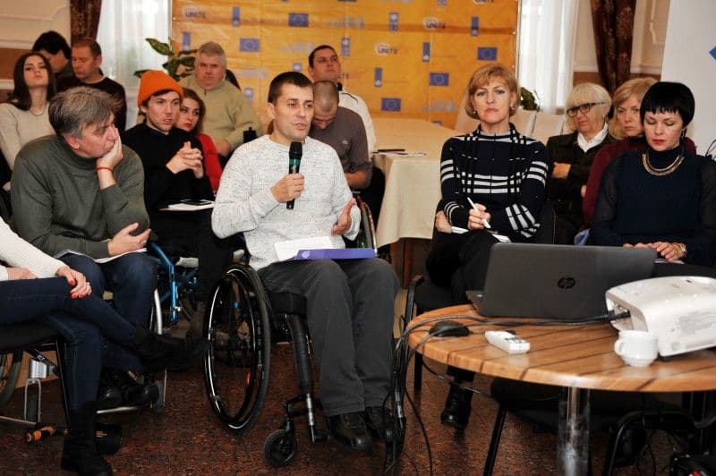 Чоловік на інвалідному візку виступає перед аудиторією, тримаючи мікрофон. Довкола учасники заходу, на фоні банер ЄС та UNITE.