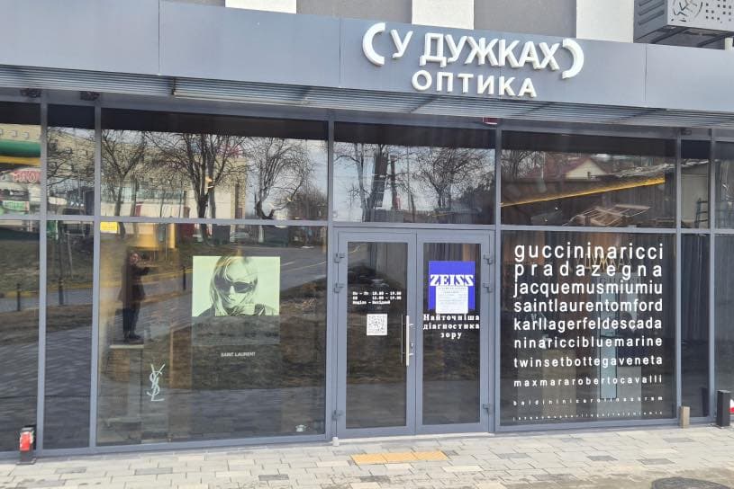 Storefront of the optical shop “У ДУЖКАХ ОПТИКА” (In Brackets Optics) with large glass doors and windows. The right window lists luxury eyewear brands including Gucci, Prada, Saint Laurent, Tom Ford, Karl Lagerfeld, Escada, Max Mara, and others. On the left window, a poster shows a person wearing sunglasses with the text “SAINT LAURENT” and the YSL logo. The door displays business hours and a ZEISS logo, indicating the use of ZEISS lenses. A Ukrainian sign reads “The most accurate vision diagnostics.” The scene highlights a modern optical store combining fashion and advanced eye care.