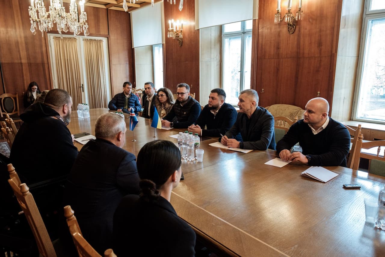 Дванадцять людей сидять за великим дерев’яним столом у конференц-залі з люстрами, дерев’яними стінами та великими вікнами. Більшість учасників — у діловому одязі, перед ними — блокноти або документи. На столі — пляшки з водою, склянки та маленький прапор України в центрі. Обстановка передає атмосферу офіційної зустрічі, присвяченої державному управлінню, політиці або плануванню, з акцентом на професіоналізм і громадську участь.