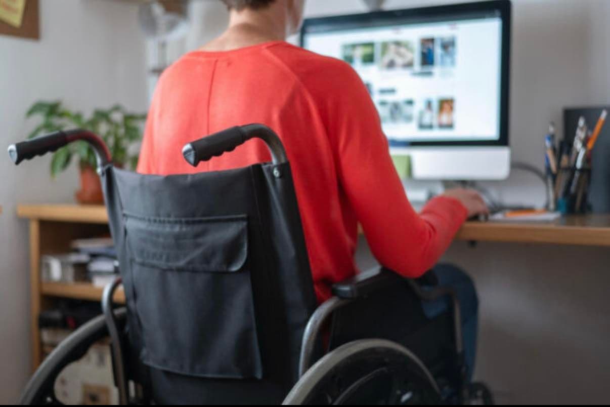 A person in a wheelchair works at a wooden desk with a computer. Wearing a red long-sleeve shirt, they sit in front of a large monitor displaying a webpage with multiple images. The desk holds office supplies, a plant, and a container with pens and pencils. In the background are shelves with books and documents. The scene emphasizes inclusivity and the integration of assistive devices in professional or remote work environments.