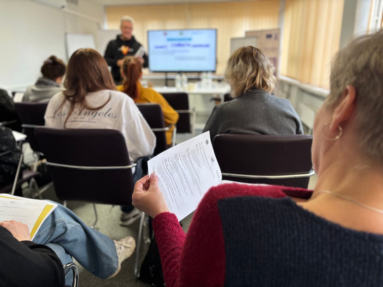 Учасники тренінгу сидять у навчальній аудиторії та переглядають роздаткові матеріали, поки тренер проводить сесію перед екраном із презентацією.»