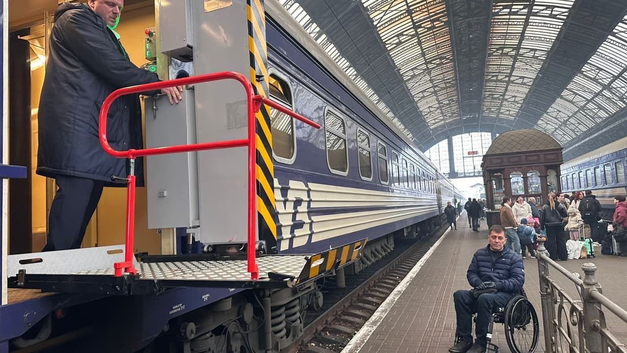 На вокзалі чоловік у темному пальті керує підйомником, що прикріплений до синього вагона поїзда з білими смугами. На платформі — чоловік на інвалідному візку та пасажири з багажем. Підйомник має червоні поручні та платформу для посадки. Сцена демонструє доступність залізничного транспорту для людей з порушеннями мобільності.