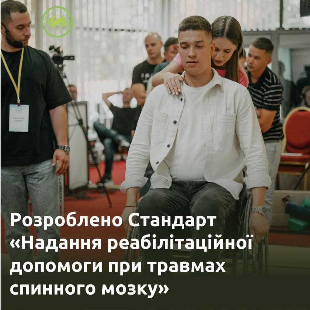 In an indoor rehabilitation setting, a young man sits in a wheelchair while a woman stands behind him, assisting or guiding him. Several other individuals are present in the background, some wearing name tags, indicating a professional or organized training session. The environment resembles a medical or conference facility. A Ukrainian-language banner in the background announces the development of a standard for providing rehabilitation assistance for spinal cord injuries.