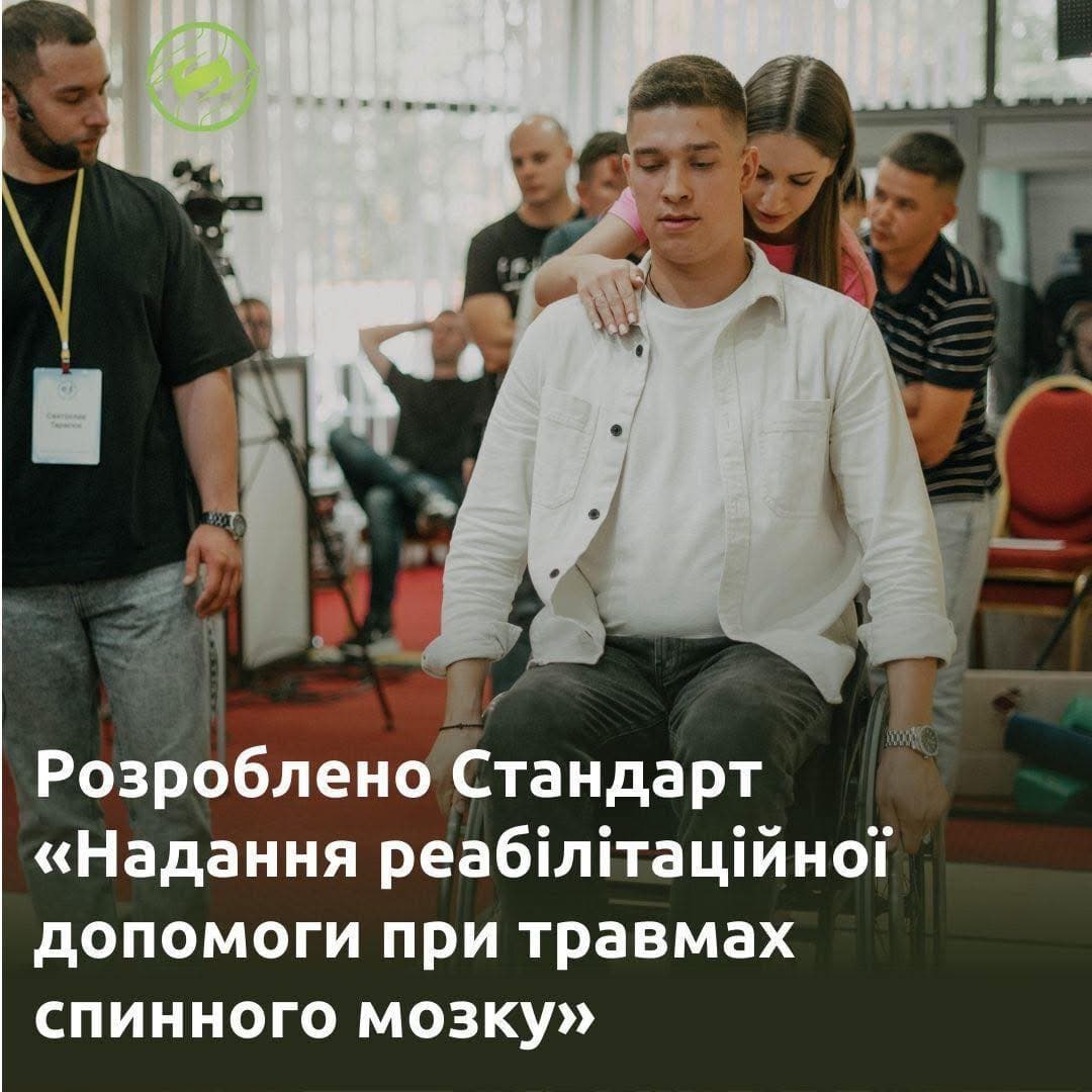 In an indoor rehabilitation setting, a young man sits in a wheelchair while a woman stands behind him, assisting or guiding him. Several other individuals are present in the background, some wearing name tags, indicating a professional or organized training session. The environment resembles a medical or conference facility. A Ukrainian-language banner in the background announces the development of a standard for providing rehabilitation assistance for spinal cord injuries.