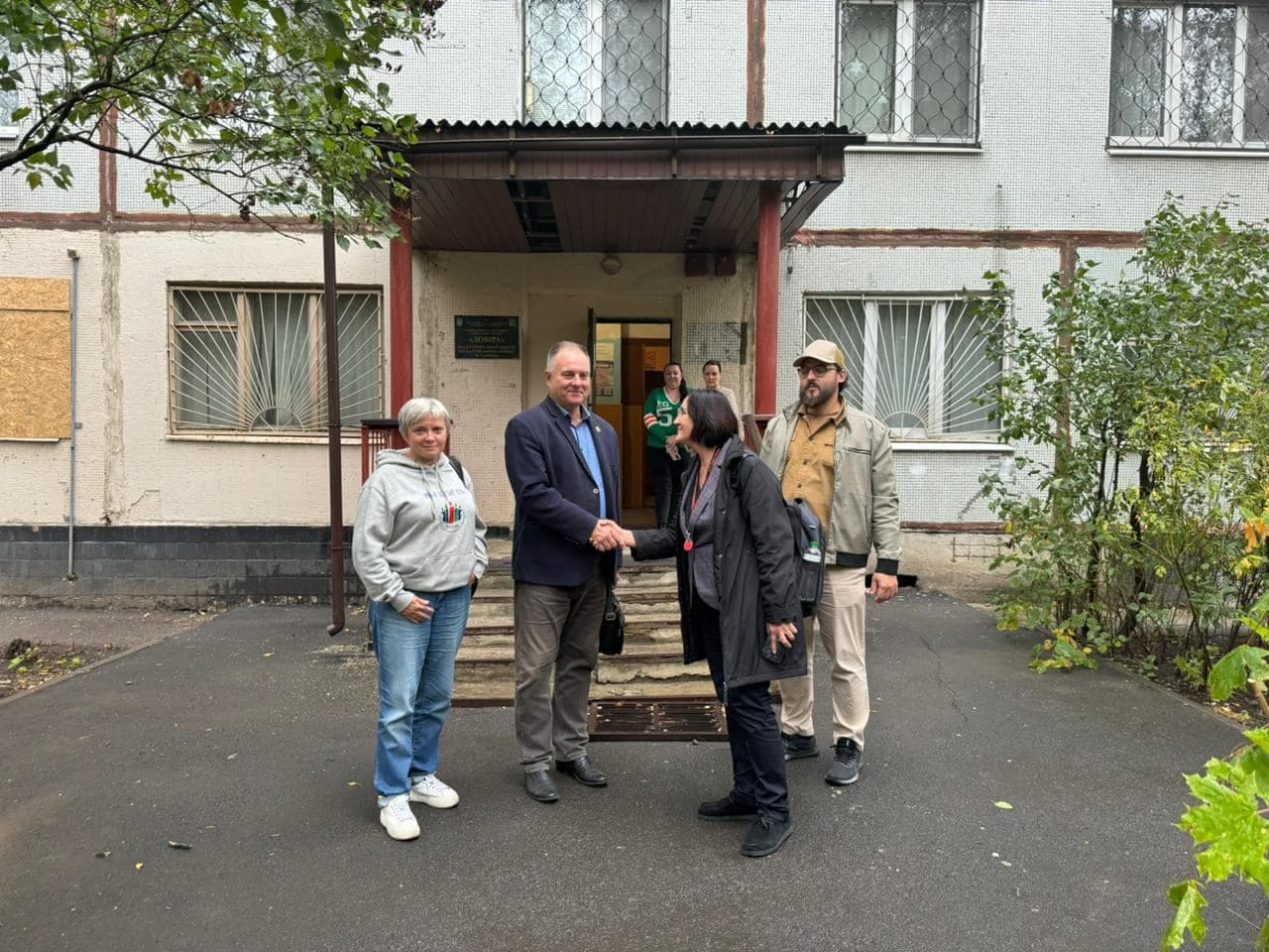 Four people are standing near the entrance of the organization's building, shaking hands. Two other people are walking out of the building.