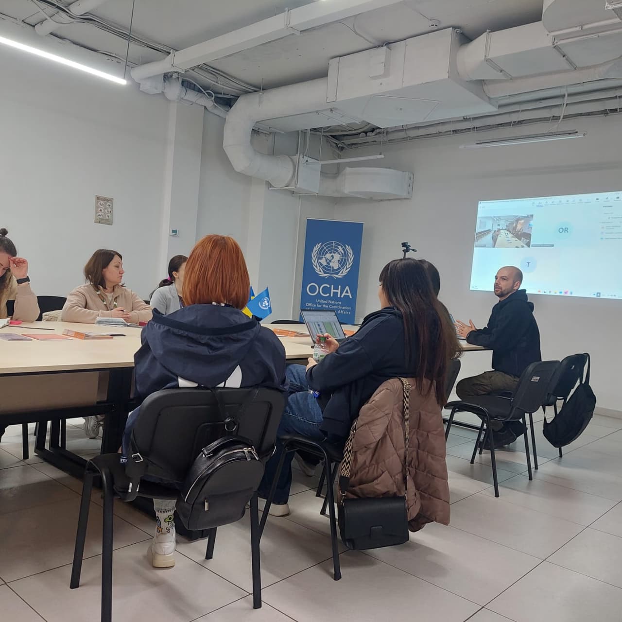 Люди в залі слухають презентацію про субсидії на оренду житла для ВПО. Дві жінки з мікрофонами сидять на сцені, за ними — екран з темою презентації.