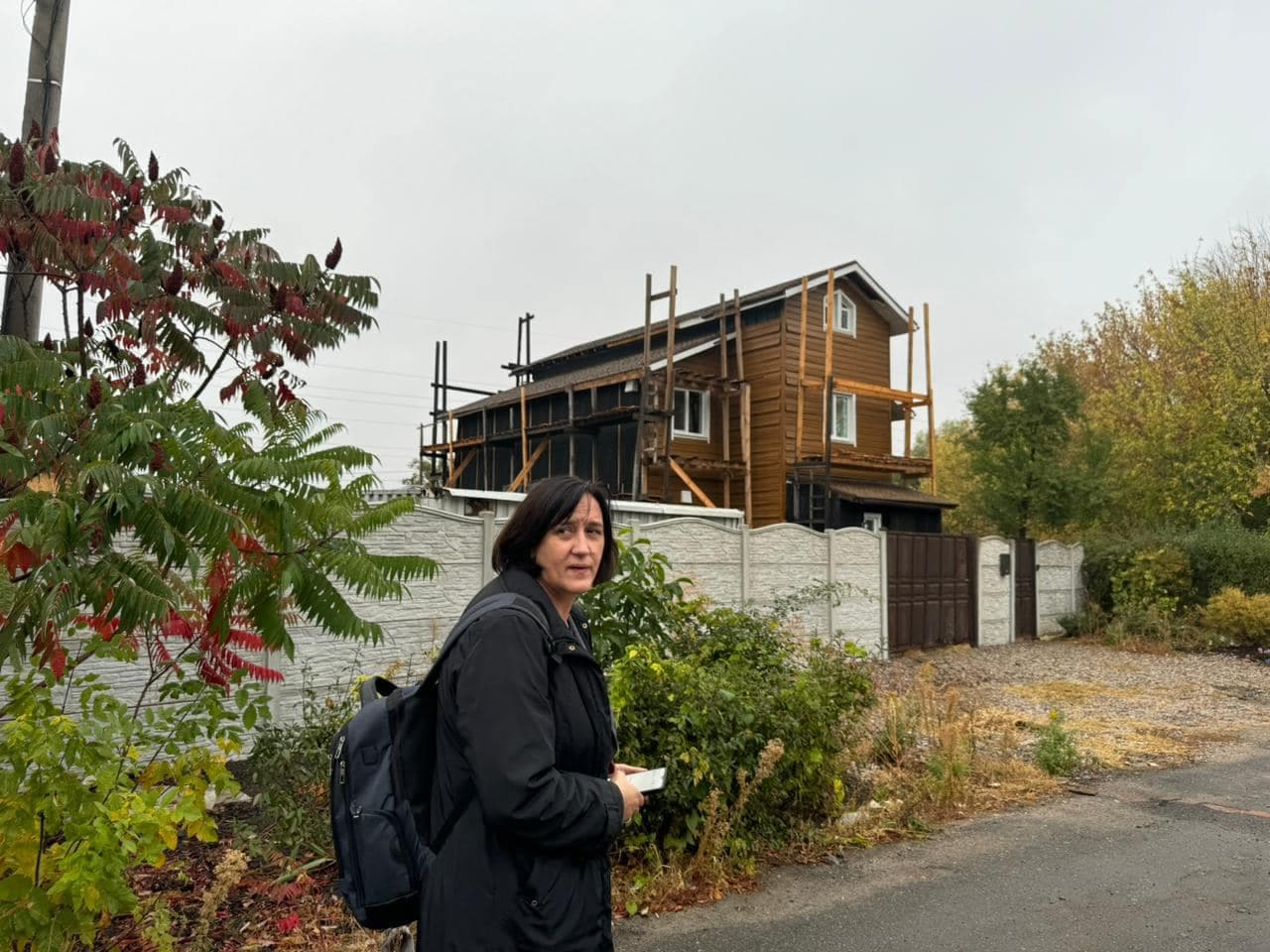 A woman stands in front of a house behind a fence, which was damaged by shelling and is currently being repaired as part of the project.