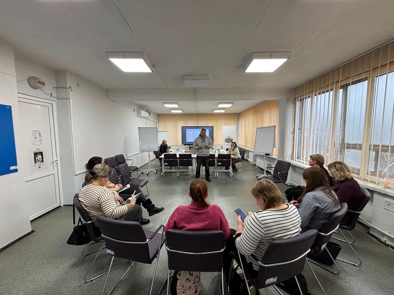 A group of people participates in a training or meeting in a conference room with white walls, large windows, and vertical blinds. Attendees sit in a semicircle of chairs, with a few seated at a table in the back. One person stands and addresses the group. Whiteboards and a screen are visible at the front. The setting is professional, focused on learning or discussion.